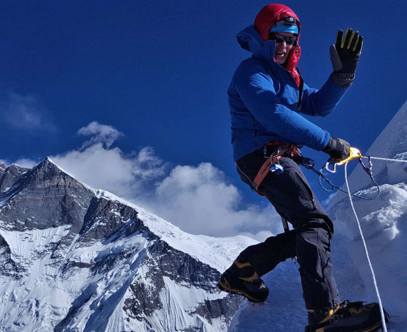 A climber waves as he begins to abseil off the summit of Island Peak. Steep mountain terrain in the distance.