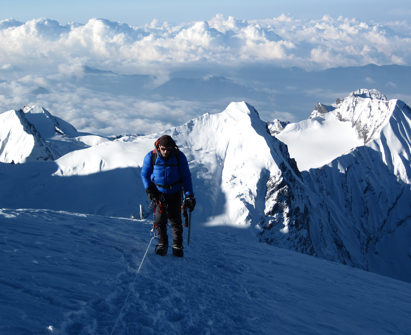 A climber uses fixed ropes as they approach the summit of Mera Peak, snow covered mountains of the Himalaya stretch into the distance behind.