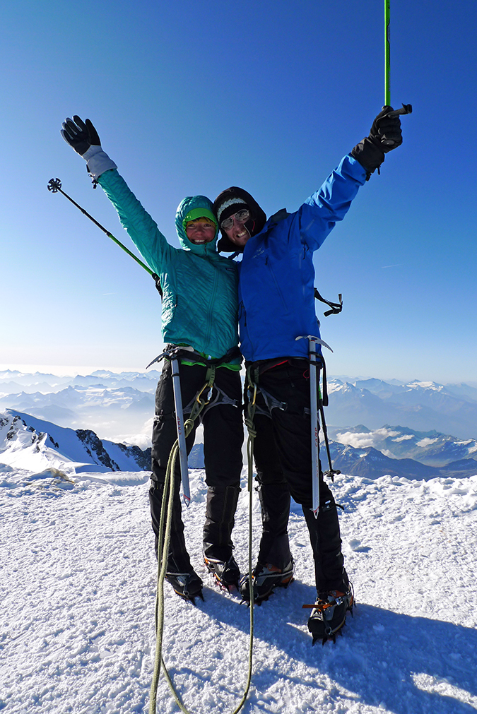 A couple stands jubilant, arms outstretched in celebration on the summit of Mont Blanc.