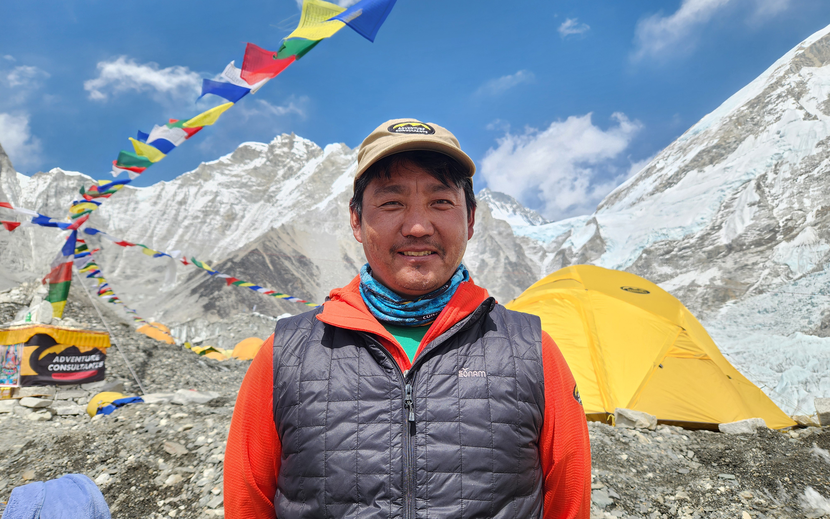 AC Trekking Sherpa Tsering Wangchu at Everest Base Camp, bright yellow tents and prayer flags wave in the breeze behind.