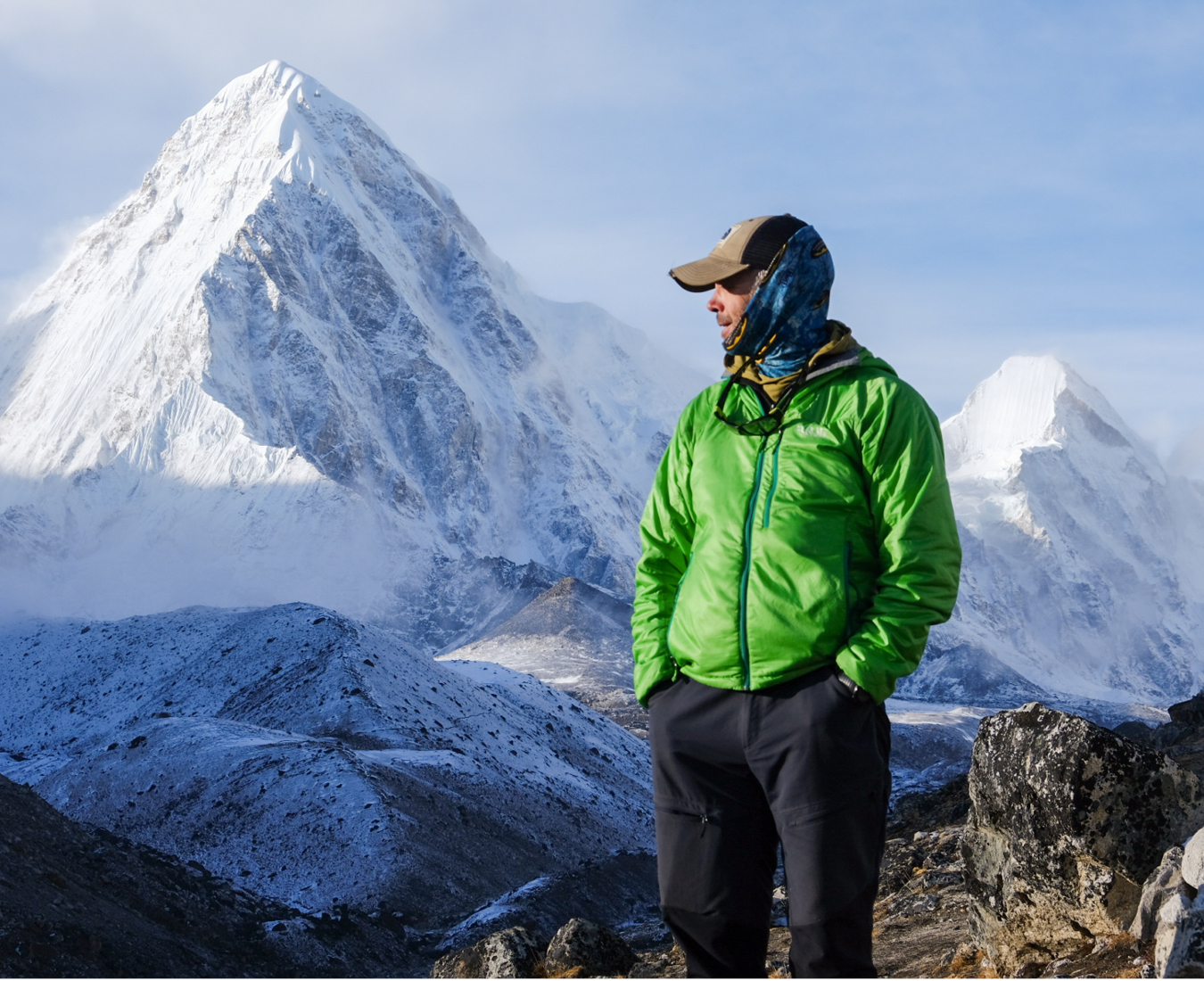 A climber in a bright green jacket contemplates the view in late afternoon light, fresh snow coating the surrounding mountains.